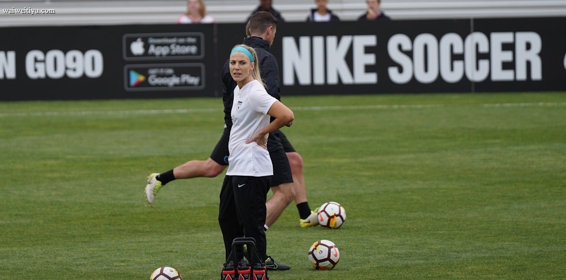 A person in sports attire stands on a soccer field, surrounded by several soccer balls and a carrier with bottles. The field is bordered by advertisements for Nike Soccer and app stores. Several people sit in bleachers in the background.