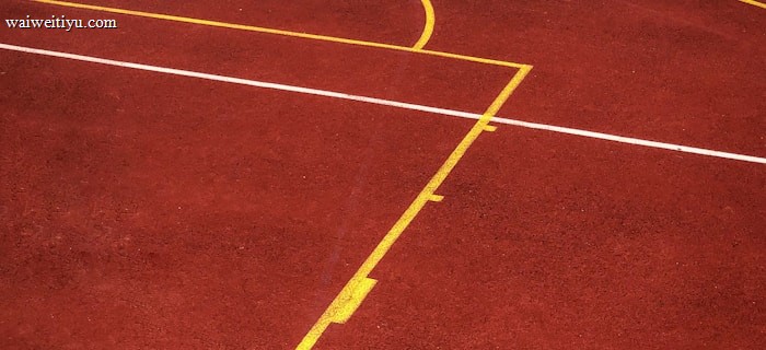 A sports court with a reddish-brown surface and bright yellow and white lines. The lines form geometric shapes, including rectangles and circles, suggesting markings for games like basketball or netball.