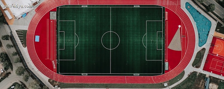 An aerial view of a large sports complex featuring a central green football field surrounded by a red running track. Adjacent to the track, there is a parking area, buildings, and pathways. Nearby, there are additional sports courts in different colors, including a blue swimming pool and orange basketball courts.