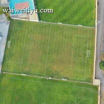 An aerial view of a sports complex featuring multiple green grass fields, including football pitches with marked goalposts. Adjacent to the fields is a blue-roofed building and a smaller area with a tennis or basketball court. Surrounding the complex, there's a road lined with trees and a dirt area with sparse vegetation.
