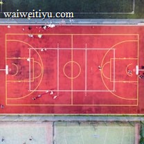 An aerial view of a sports court with a vibrant red surface and yellow lines marking different sections for basketball and soccer. Several people are present on the court, some engaged in activities, while others are standing along the perimeter. The court is enclosed by a metal fence and surrounded by grass on all sides.