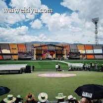 A sports stadium with a large stage set up on the field, hosting an outdoor event. Spectators wearing colorful hats sit in the foreground. The seating areas display vibrant colors, and a clear blue sky with scattered clouds is visible. Large screens display graphics related to the event.