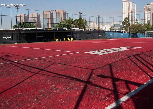 A sports court with a red playing surface and white lines, surrounded by a black mesh fence. In the background, tall buildings are visible, along with some trees and a few yellow chairs positioned near the fence.