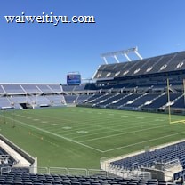 A large, empty sports stadium with blue seats surrounding a well-maintained green grass field. Bright sunlight creates clear shadows, and there are two goal posts on opposite ends of the field. A big electronic scoreboard displays some graphics, and the sky is clear and blue.